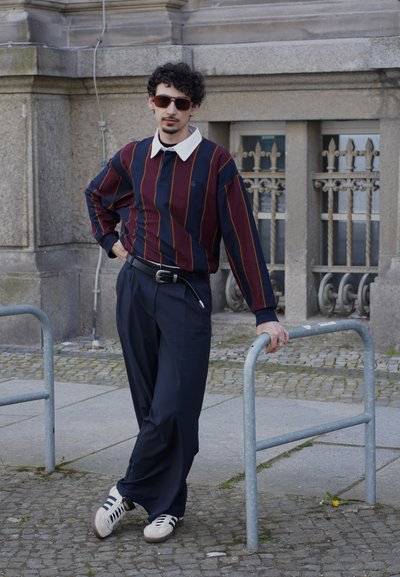 Polo de manga larga a rayas en rojo oscuro y azul marino con cuello blanco, combinado con pantalones azul marino y zapatillas crema, apoyado en una barandilla de metal.