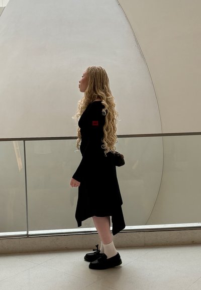Vestido negro de manga larga con un parche rojo; cabello texturizado en ondas sueltas; calcetines blancos y zapatos negros; de pie contra un fondo claro y curvado.