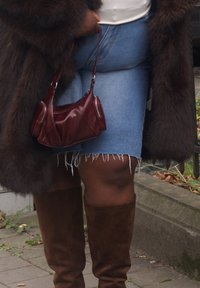 Personne portant un manteau en fourrure marron, une jupe en jean et des bottes marron montant jusqu'aux genoux, tenant un petit sac à main en cuir bordeaux par la bandoulière.