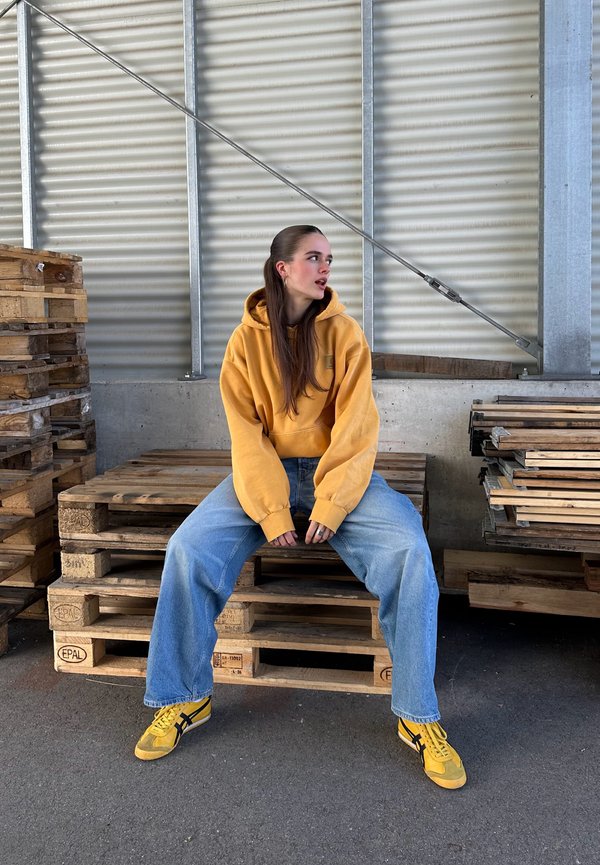 Sudadera amarilla con mangas voluminosas, combinada con jeans azules de gran tamaño y zapatillas amarillas. Palets de madera en el fondo.