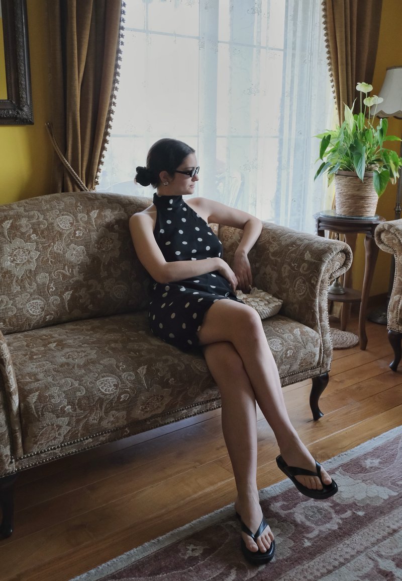Femme en robe noire à pois et lunettes de soleil assise jambes croisées sur un canapé à motifs dans une pièce ensoleillée avec des rideaux et des plantes.