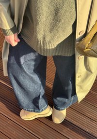 Personne portant des bottes beige, un jean large bleu foncé, un pull en tricot gris, un manteau beige, et tenant un sac à main tressé marron, debout sur une terrasse en bois.