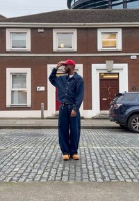 Hombre con chaqueta y pantalones de mezclilla oscuros, gorra roja y zapatos beige de pie en una calle empedrada frente a un edificio de ladrillo con ventanas y puerta con marcos blancos.