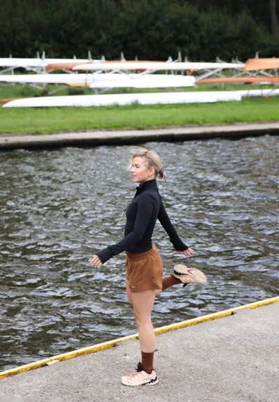 Atleta con una camiseta de manga larga negra y pantalones cortos marrones, realizando un estiramiento de pierna en un paseo marítimo, con zapatos rosas y calcetines marrones.