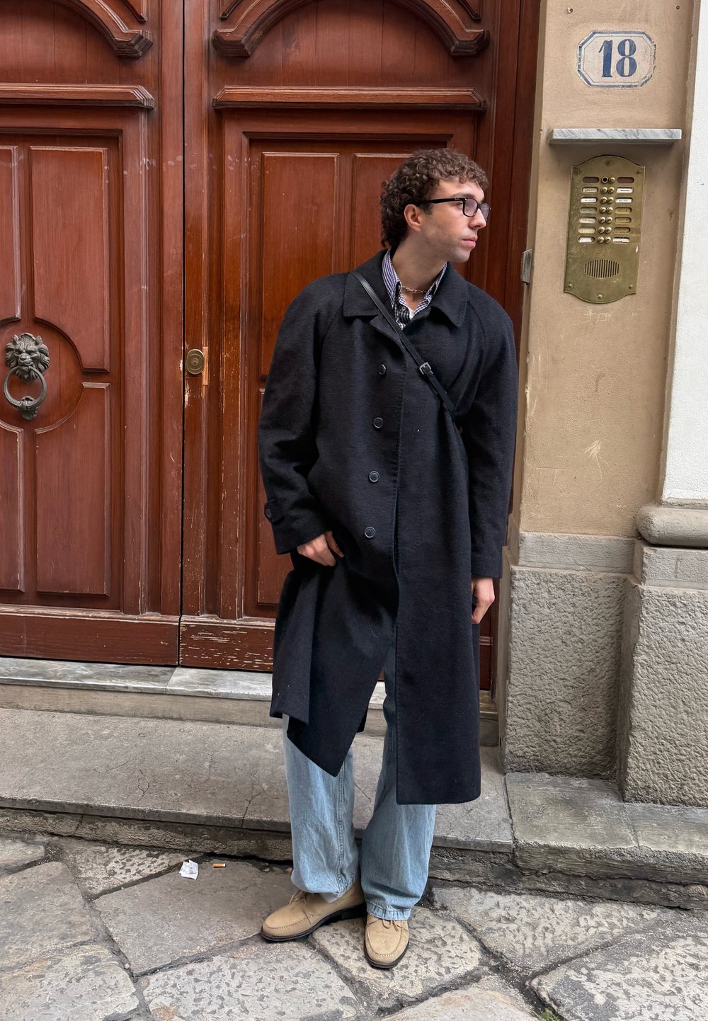 Homme portant un manteau noir, un jean clair et des chaussures beiges, debout près d'une grande porte en bois avec un interphone en laiton et le numéro 18 sur le mur.
