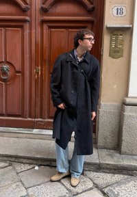 Homme portant un manteau noir, un jean clair et des chaussures beiges, debout près d'une grande porte en bois avec un interphone en laiton et le numéro 18 sur le mur.