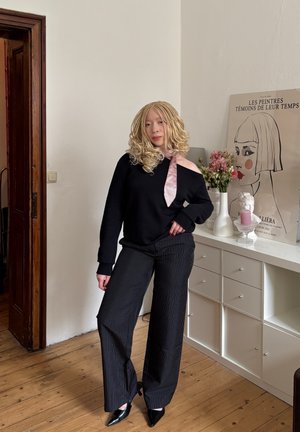 Sudadera negra sin hombros con un detalle de lazo en rosa claro, combinada con pantalones de pierna ancha a rayas negras y tacones de punta negra.