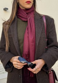 Ruskea herringbone-takki oliivi vihreän paidan päällä, viininpunainen huivi frangeilla, mustat aurinkolasit käsissä, hopearenkaita sormissa, olkalaukku näkyvillä.