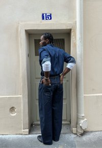 Homme aux cheveux tressés portant un jean foncé, debout les mains dans les poches arrière près d'une porte beige portant le numéro 15 dans une rue étroite.