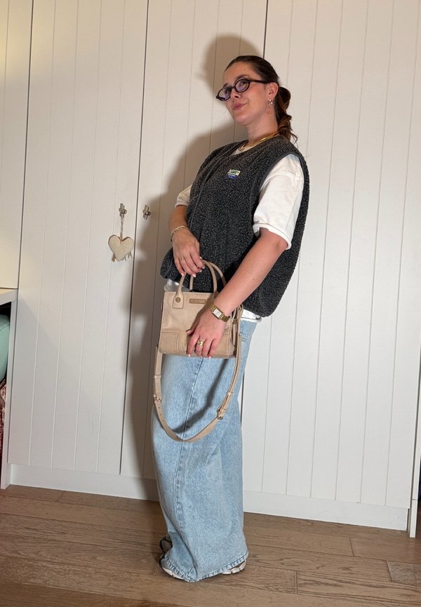 Femme portant des lunettes, t-shirt blanc, gilet gris foncé, jean large bleu clair, tenant un sac à main beige, debout à l'intérieur contre un mur blanc à panneaux.
