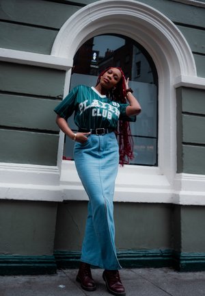 Camisa verde a rayas verticales con el texto "PLAYER CLUB", combinada con una falda de mezclilla azul clara con una abertura frontal y botas marrones con cordones.