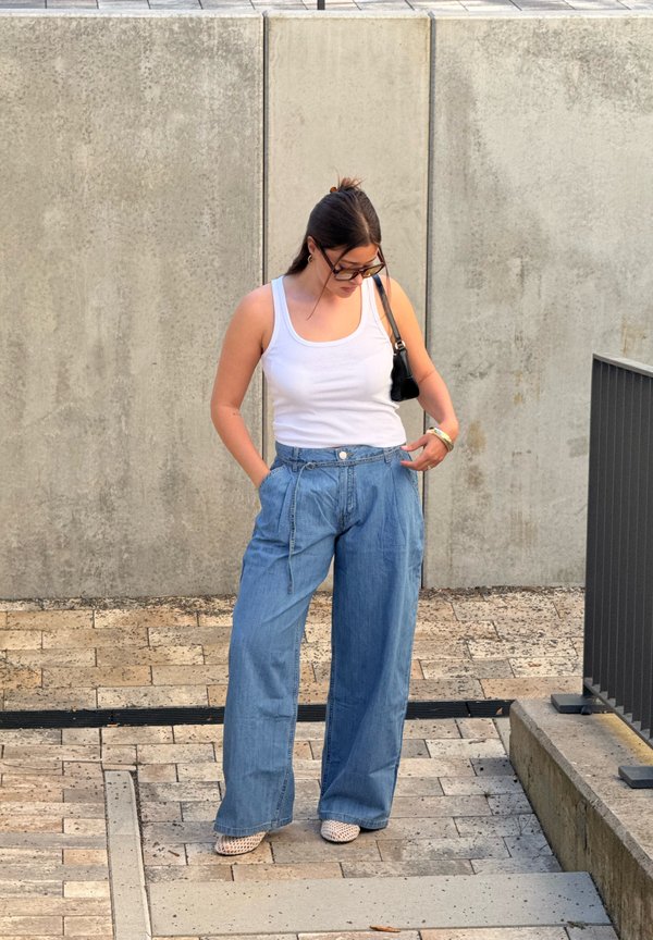 Femme portant un débardeur blanc, un jean bleu à jambes larges, des lunettes de soleil et un sac épaule noir, debout sur des marches en pierre à l'extérieur.