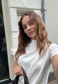 Hvit topp med draperinger og høy hals, kombinert med en svart veske. Myk tekstur og flytende design, med løs, bølget brunt hår.
