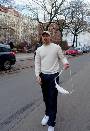 Suéter beige, pantalones azul marino, zapatillas blancas. Sosteniendo un bolso blanco de hombro con correa ajustable. Escena al aire libre con árboles y edificios.
