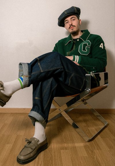 Chaqueta varsity verde con acentos blancos, gorra azul, jeans de mezclilla oversized con dobladillos enrollados, calcetines blancos con rayas de colores y mocasines grises.
