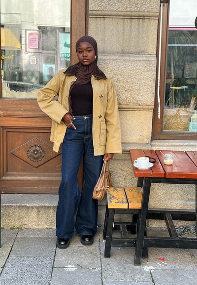 Chaqueta caqui con cuello marrón, top negro ajustado, vaqueros azules de cintura alta y zapatos negros; sosteniendo un bolso marrón; mesa de madera cercana con una taza.