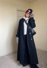 Long manteau bleu marine à double boutonnage, pull gris, pantalon bleu marine, sac à bandoulière noir, gants gris et bonnet rayé avec un logo.