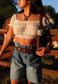 White cropped blouse with textured fabric, high-waisted denim shorts, a brown belt with metal accents, and a brown bag with studs and a floral detail.