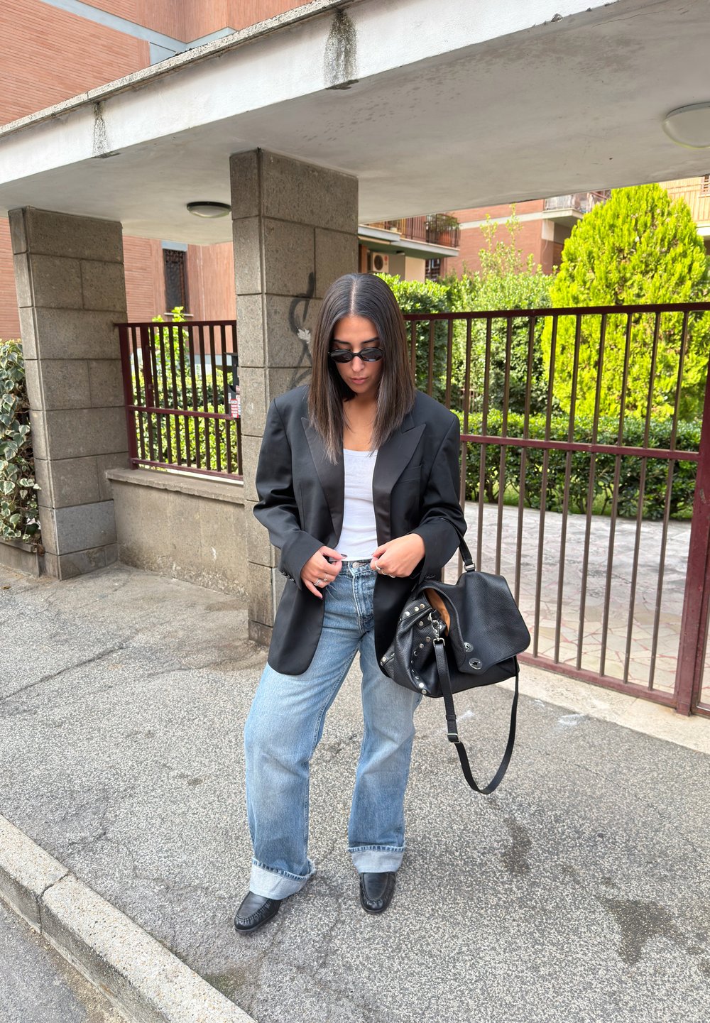 Czarna oversize'owa marynarka, biała koszulka na ramiączkach, jasnoniebieskie szerokie spodnie jeansowe, czarna skórzana torebka i czarne buty, na tle kamieni i zieleni.
