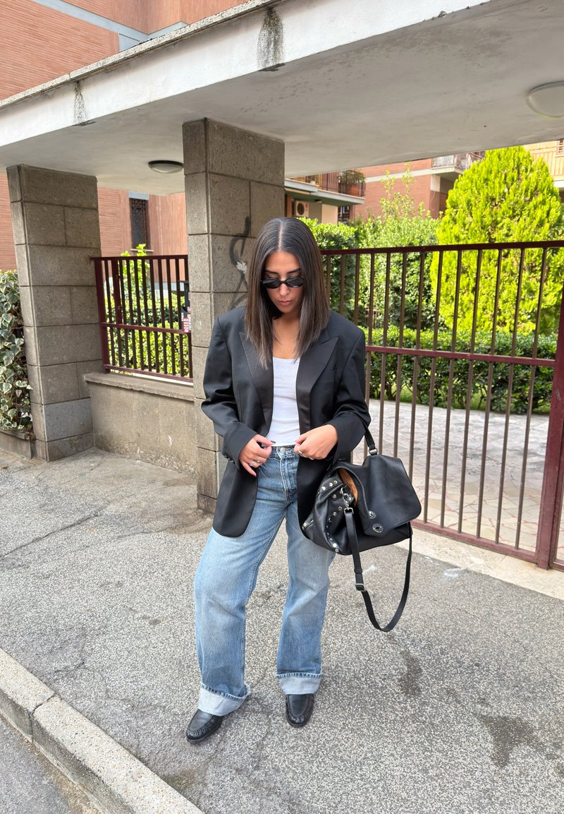 Blazer preto oversize, top branco de alças, jeans azul claro de perna larga, mala de couro preta e sapatos pretos, com fundo de pedra e vegetação.