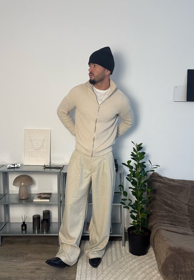 Hombre con suéter beige, pantalones crema, gorro negro, de pie en la sala cerca de una planta, una estantería con decoración y un sofá marrón.