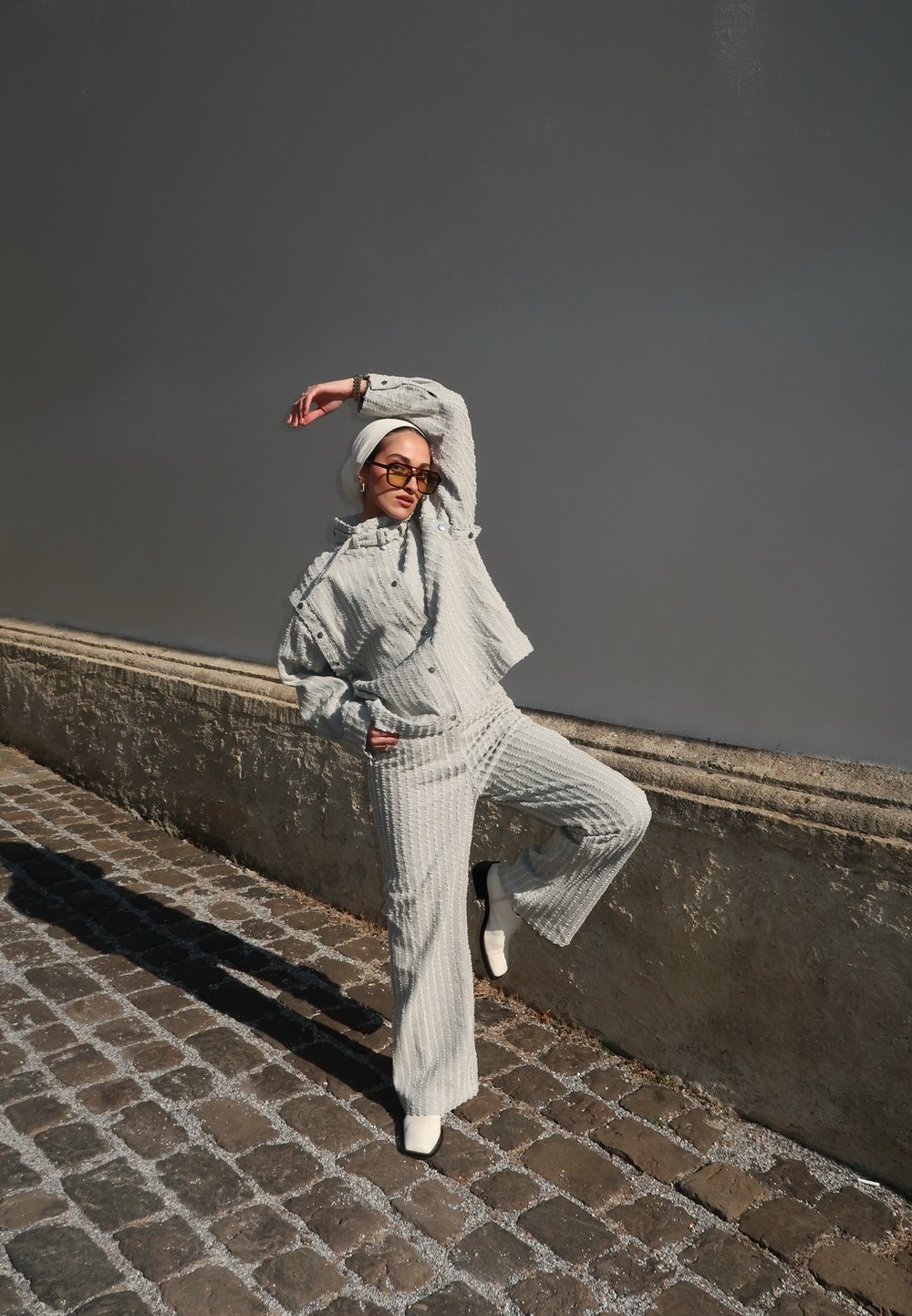 Outfit grigio strutturato con giacca corta e pantaloni a campana, abbinato a stivali bianchi alla caviglia e occhiali da sole, su uno sfondo neutro.