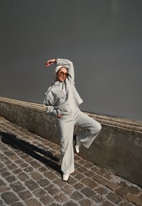 Outfit grigio strutturato con giacca corta e pantaloni a campana, abbinato a stivali bianchi alla caviglia e occhiali da sole, su uno sfondo neutro.