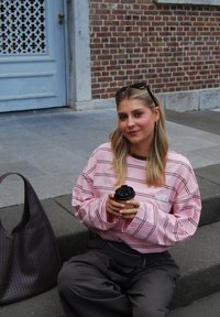 Hellrosa übergroßer Pullover mit dunklen Streifen, schwarze Hose, hält einen To-Go-Becher. Braune gewebte Tasche in der Nähe, sitzt auf Steinstufen.