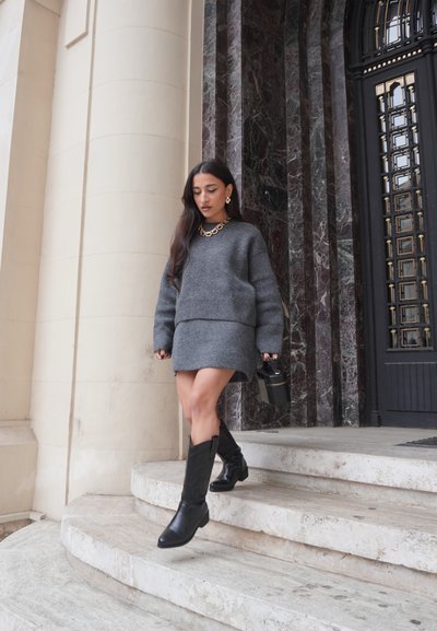 Suéter gris de punto y falda a juego, botas de cuero negras hasta la rodilla, collar de cadena dorada, bajando escaleras de mármol, bolso minimalista.