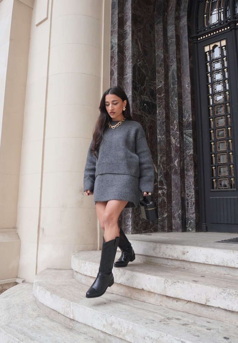 Grauer Strickpullover und passender Rock, kniehohe schwarze Lederstiefel, goldene Kettenhalskette, gehend die Marmortreppe hinunter, minimalistische Tasche.