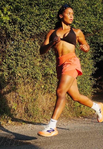 Figura atlética con un top corto negro y pantalones cortos naranjas, corriendo por un sendero. Lleva calcetines blancos y zapatillas naranjas con acentos morados.