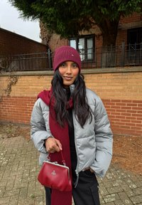 Siva puhasta jakna, bordo rebrasta pletena kapa, usklađena šal, in rdeča torba iz umetnega usnja s srebrnimi dodatki, postavljena ob zid iz opeke.