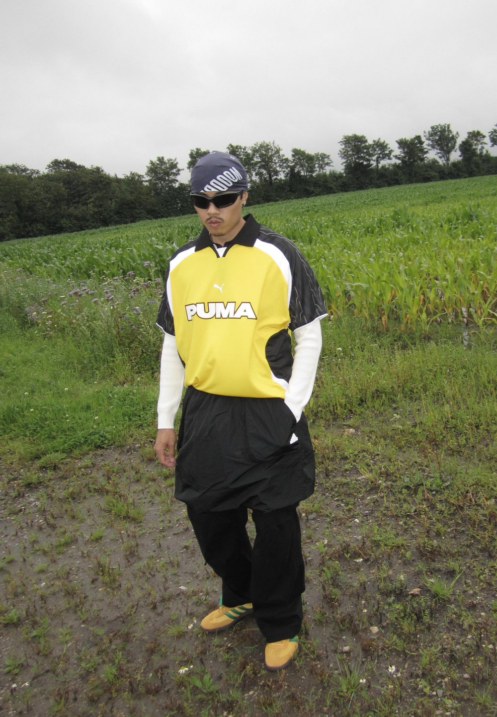 Black and yellow Puma jersey with white accents, baggy black shorts, and yellow shoes featuring green stripes; standing on grass in a field.