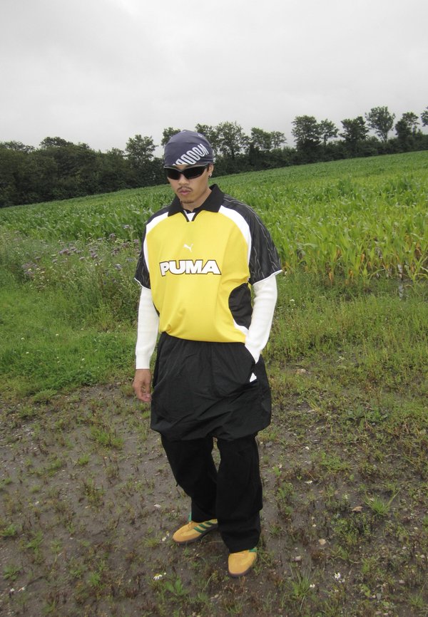 Black and yellow Puma jersey with white accents, baggy black shorts, and yellow shoes featuring green stripes; standing on grass in a field.