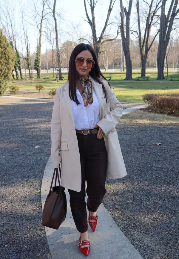 Femme portant un blazer beige, une chemise blanche, un pantalon sombre, des chaussures pointues rouges, des lunettes de soleil et une écharpe, marchant sur un chemin de parc en portant un sac à main marron.