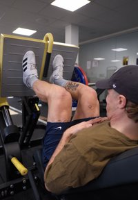 Máquina de prensa de piernas con una plataforma gris y detalles amarillos. Una persona con zapatos grises y pantalones cortos azules realiza el ejercicio, mostrando tatuajes en las piernas.