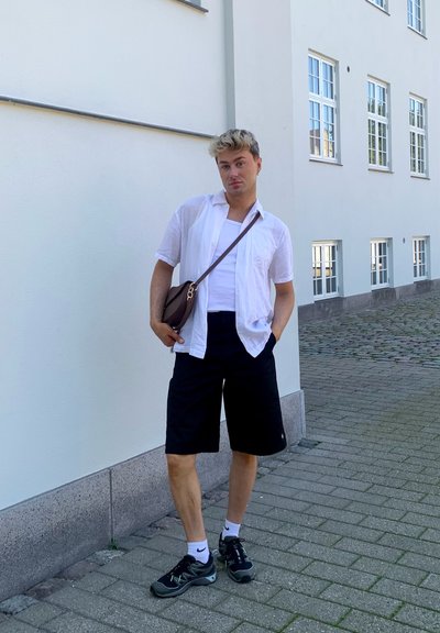 Camisa blanca de manga corta ligera, pantalones cortos negros, zapatillas oscuras con calcetines blancos, bolso cruzado marrón, de pie contra una pared de color claro.