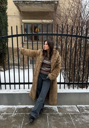 Joven con un abrigo largo de piel marrón y un suéter a rayas apoya en una valla de metal negro con nieve en el suelo y arbustos sin hojas detrás.