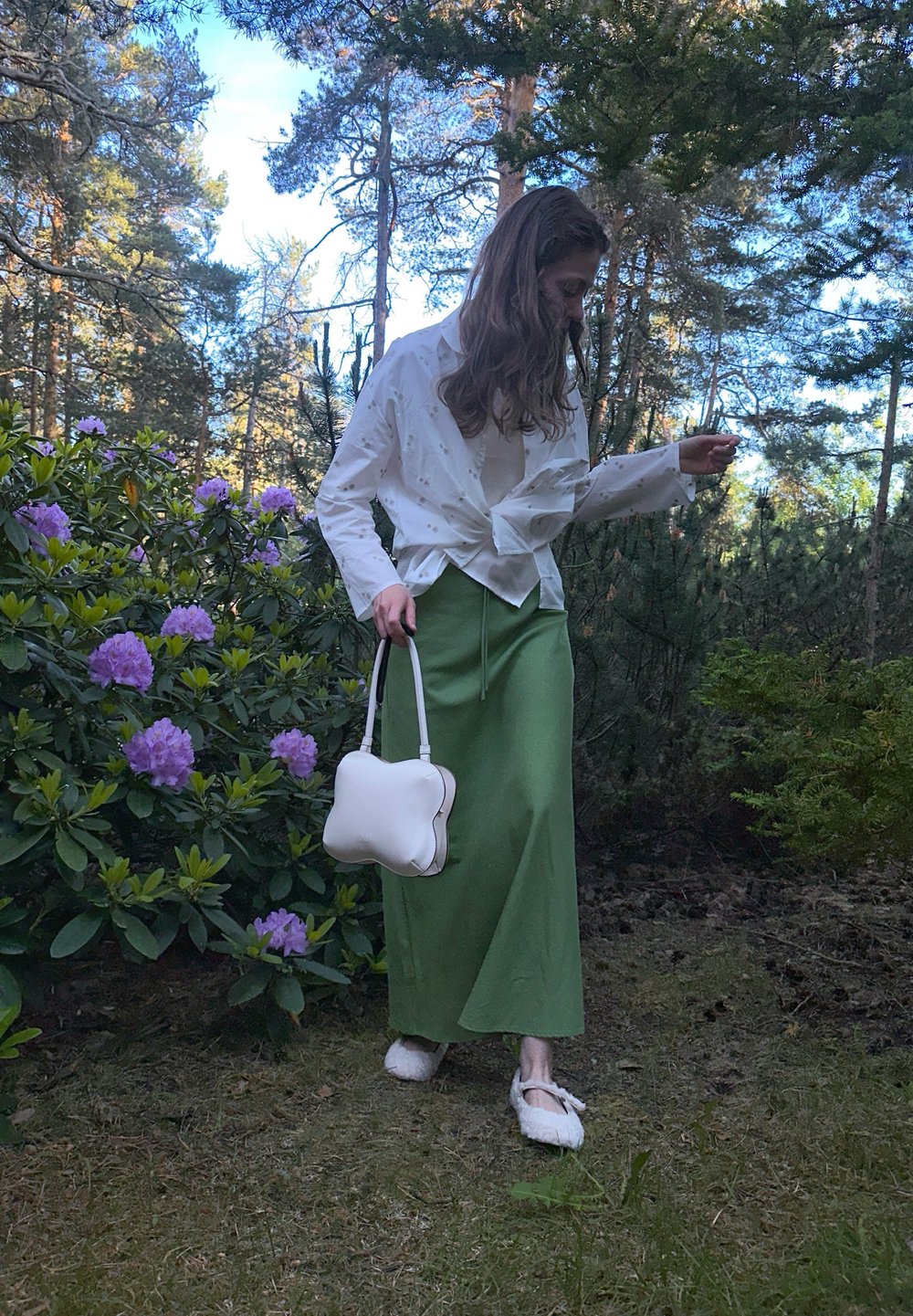 Chemise blanche à manches longues avec un nœud sur le devant, jupe maxi verte, sac à main en forme d'os blanc et chaussons blancs duveteux dans un cadre naturel avec de la verdure.