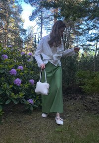 Chemise blanche à manches longues avec un nœud sur le devant, jupe maxi verte, sac à main en forme d'os blanc et chaussons blancs duveteux dans un cadre naturel avec de la verdure.