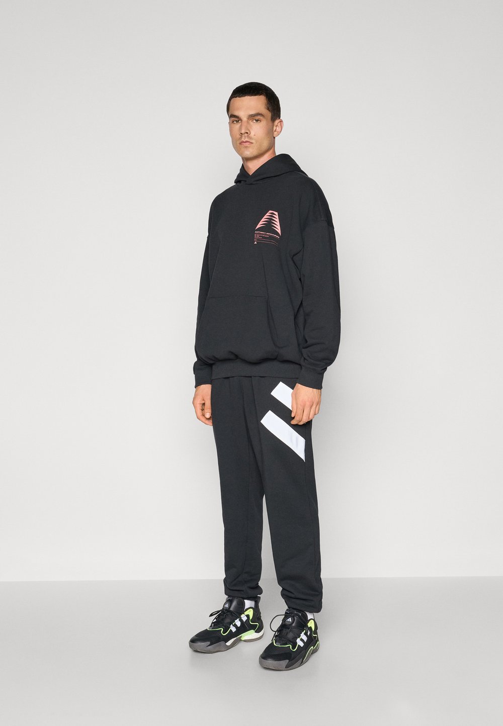 Melna kapucēta džemperis ar rozā grafiku, apvienots ar melniem sporta biksēm ar baltām diagonālām svītrām. Melni kedu ar zaļiem akcentiem.