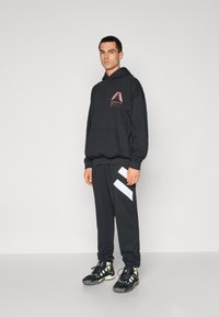 Melna kapucēta džemperis ar rozā grafiku, apvienots ar melniem sporta biksēm ar baltām diagonālām svītrām. Melni kedu ar zaļiem akcentiem.