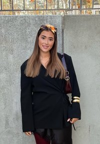 Melna, garām piedurknēm žakete ar aptinam dizainu un sasienamu jostu, kombinēta ar bordo krāsas biksēm. Komplektā zelta aproces akcenti un tumša plecu soma.