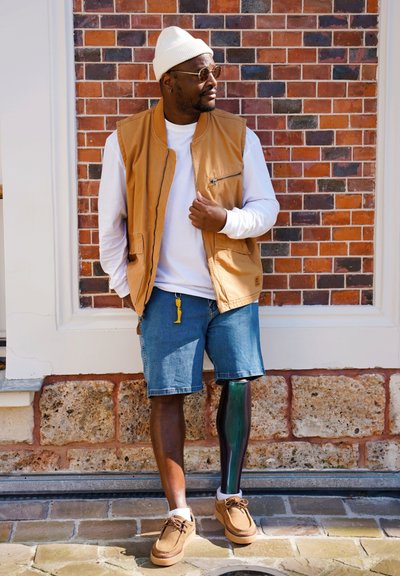 Un chaleco sin mangas color tostado sobre una camisa blanca de manga larga, combinado con pantalones cortos de mezclilla azul. Se pueden ver zapatos marrones y una pierna prostética verde.