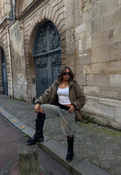 Joven mujer con gafas de sol, chaqueta de piel, top blanco, falda larga y botas negras posa en una calle adoquinada junto a una puerta gris ornamentada y una pared de piedra.