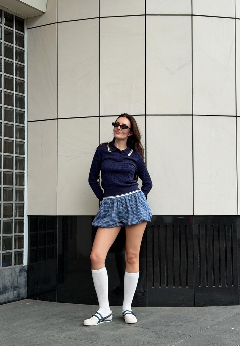 Donna con maglione scuro, gonna blu a quadri, calze bianche al ginocchio e occhiali da sole, in piedi contro una parete piastrellata con le mani dietro la schiena.