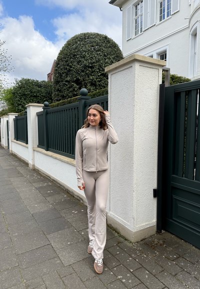 Chaqueta beige claro con cremallera y pantalones ajustados a juego, combinados con zapatillas marrones; tejido suave y diseño elegante, en un paseo pavimentado.