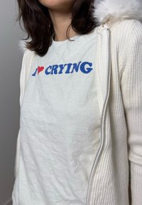 Fehér cipzáras kapucnis pulóver bordázott textúrával és műszőrme szegéllyel a kapucnin, világosszürke póló fölött, amelyen kék és piros felirat áll: "I ❤️ CRYING."