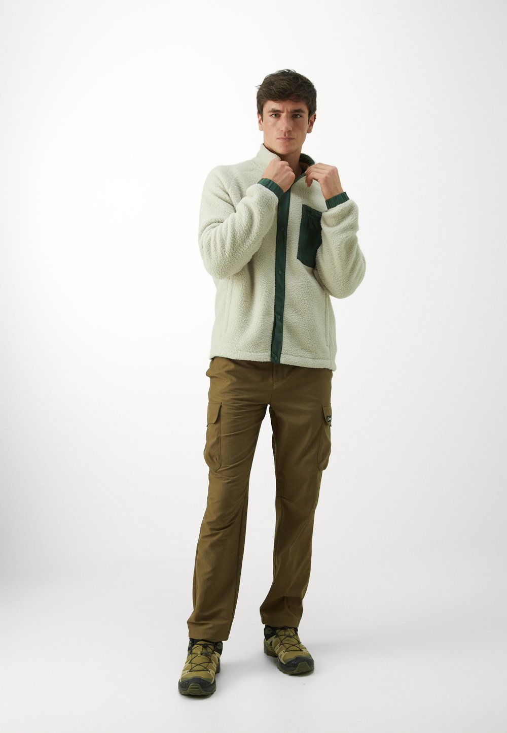 Kreminė flisinė striukė su tamsiai žaliais mygtukais ir kišene, derinama su rudomis kargo kelnėmis ir pilkomis lauko bateliais.