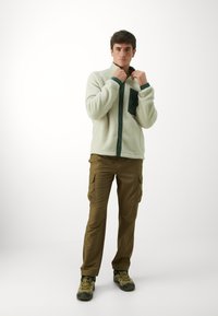 Kreminė flisinė striukė su tamsiai žaliais mygtukais ir kišene, derinama su rudomis kargo kelnėmis ir pilkomis lauko bateliais.